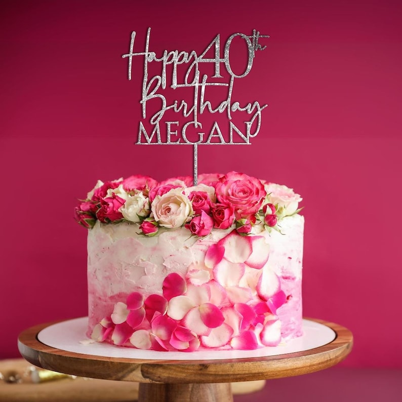 May include: Silver glitter cake topper with the text "Happy 40th Birthday MEGAN". The topper is on a pink and white frosted cake decorated with pink rose petals and pink and white roses.