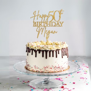May include: Gold glitter cake topper with the text "Happy 50th Birthday Megan". The topper is on a white cake with chocolate frosting and sprinkles.
