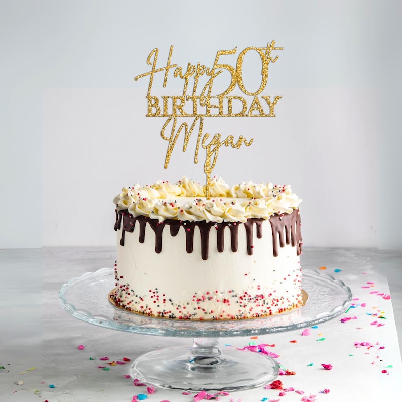 May include: Gold glitter cake topper with the text "Happy 50th Birthday Megan". The topper is on a white cake with chocolate frosting and sprinkles.
