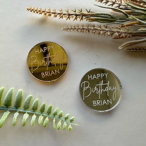 May include: Two round, mirrored acrylic birthday decorations with the text "Happy Birthday Brian" in white lettering. One decoration is gold and the other is silver.