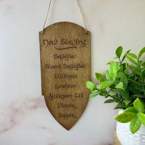 May include: Wooden sign with the words "Now Serving" and a list of meal times, including "Breakfast," "Second Breakfast," and "Dinner." The sign is shield-shaped and hangs from a natural twine string.