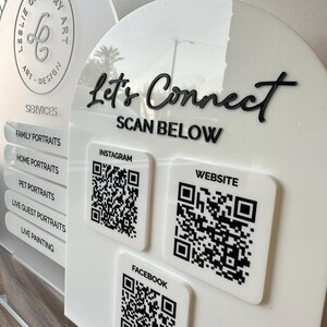 Op de afbeelding: Wit acryl bord met zwarte tekst "Let's Connect" en "Scan Below". QR-codes voor Instagram, Website en Facebook worden weergegeven. Het bord vermeldt ook portretservices.