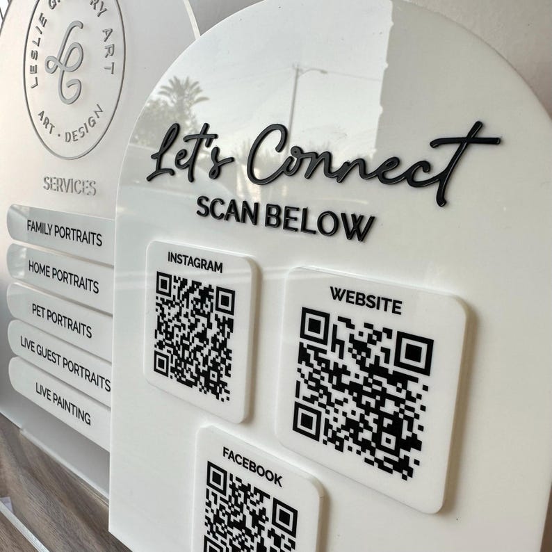 Op de afbeelding: Wit acryl bord met zwarte tekst "Let's Connect" en "Scan Below". QR-codes voor Instagram, Website en Facebook worden weergegeven. Het bord vermeldt ook portretservices.