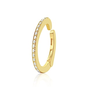 May include: A gold-colored ear cuff with a row of small, clear gemstones along the outer edge. The cuff has a simple, modern design and a hinged clasp for easy wear. The background is white.
