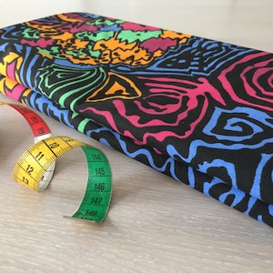 May include: A folded fabric with a colorful abstract pattern in shades of blue, pink, green, orange, and yellow on a black background. A yellow and green measuring tape is next to the fabric.