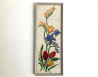 Puede incluir: Un bordado enmarcado con un arreglo floral. La obra presenta una variedad de flores en tonos rojos, amarillos, naranjas y azules, con hojas verdes. Las flores están dispuestas sobre un fondo beige, y el marco es de color gris claro.