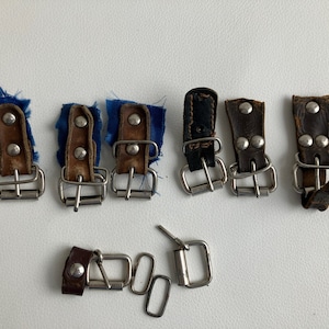Könnte beinhalten: Ein Satz von sieben Metallschnallen mit Lederriemen. Die Schnallen sind silberfarben und die Riemen sind braun und blau.