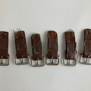 Könnte beinhalten: Sechs braune Ledergürtelschlaufen mit silbernen Schnallen. Die Schlaufen sind abgenutzt und haben sichtbare Nähte.