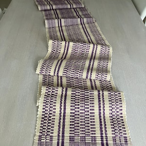 May include: A long, narrow table runner with a woven pattern of purple and cream stripes. The runner is made of a soft, textured fabric and has a subtle sheen.