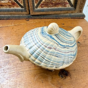 May include: A ceramic teapot with a shell-like design in shades of blue, cream, and brown. The teapot has a spout, handle, and a small knob on the lid. The teapot is placed on a wooden surface.