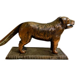 May include: A bronze-coloured dog-shaped nutcracker. The dog stands on a rectangular base with a lever extending from its back. The nutcracker has a detailed, textured surface and open mouth.