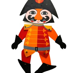 May include: A vintage wooden toy soldier with a black hat, orange face, and a red and orange uniform. The toy has black boots and gloves, and a decorative design. The toy is a classic, colorful piece.