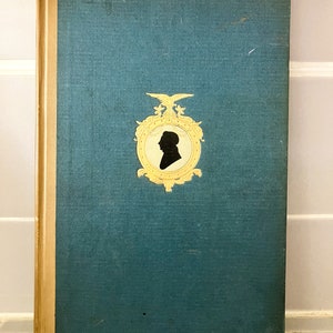 Op de afbeelding: Oud boek met een blauwgroene kaft en gouden details. De kaft heeft een gouden embleem met een zwart silhouet van een persoonsprofiel. De rug is lichtbeige van kleur. Het boek is in goede staat.