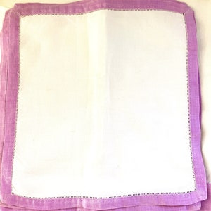 May include: A stack of white linen napkins with a lavender border. Each napkin has a stitched edge detail. The napkins are folded and stacked, showing the contrast between the white center and the colored border.