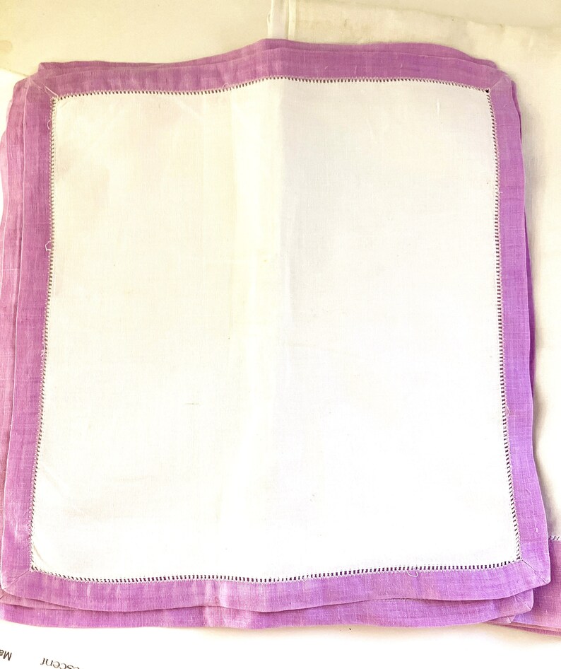 May include: A stack of white linen napkins with a lavender border. Each napkin has a stitched edge detail. The napkins are folded and stacked, showing the contrast between the white center and the colored border.