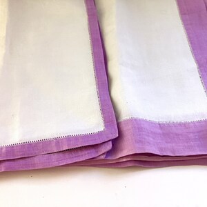 May include: Two white linen napkins with a lavender border. The napkins are folded and stacked, showcasing the contrast between the white center and the lavender edges. The edges are finished with a delicate stitched detail.