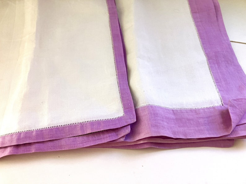 May include: Two white linen napkins with a lavender border. The napkins are folded and stacked, showcasing the contrast between the white center and the lavender edges. The edges are finished with a delicate stitched detail.