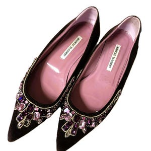 Puede incluir: Un par de bailarinas de ante marrón oscuro con punta. Los zapatos presentan una disposición decorativa de piedras preciosas moradas y rosas alrededor de la puntera. La plantilla interior es rosa y la marca "MANOLO BLAHNIK" está impresa.