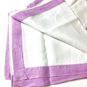 May include: White linen napkins with a lavender border. The napkins are folded and stacked, showcasing the contrasting colors and textures. The lavender border is a solid color, and the napkins have a simple, elegant design.