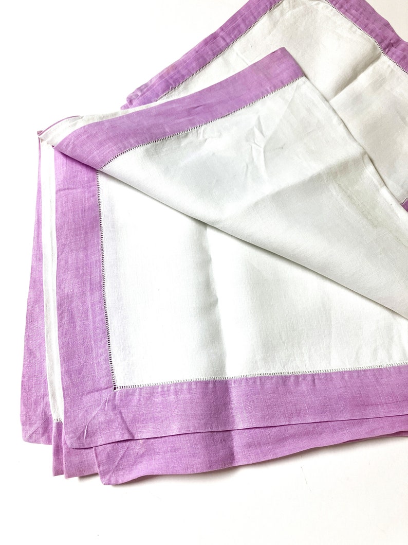 May include: White linen napkins with a lavender border. The napkins are folded and stacked, showcasing the contrasting colors and textures. The lavender border is a solid color, and the napkins have a simple, elegant design.