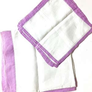 May include: Two white linen napkins with a lavender border and a decorative stitch. One napkin is folded, and the other is partially unfolded, displaying the contrast between the white center and the colored edges. The napkins are on a white background.