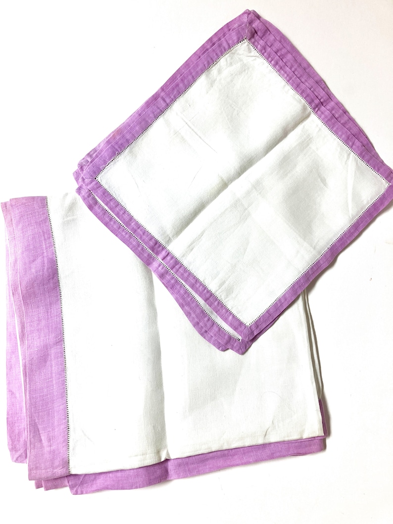 May include: Two white linen napkins with a lavender border and a decorative stitch. One napkin is folded, and the other is partially unfolded, displaying the contrast between the white center and the colored edges. The napkins are on a white background.