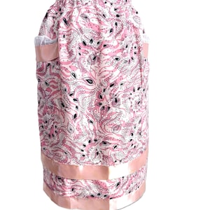 May include: A pink and white patterned skirt with a paisley design. The skirt has a satin ribbon trim at the hem and around the pockets. The skirt is displayed on a wooden mannequin.