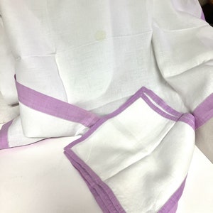 May include: White linen tablecloth with a lavender border and matching napkins. The tablecloth is folded, showing the lavender trim and a corner of a folded napkin. The fabric has a slightly textured appearance.