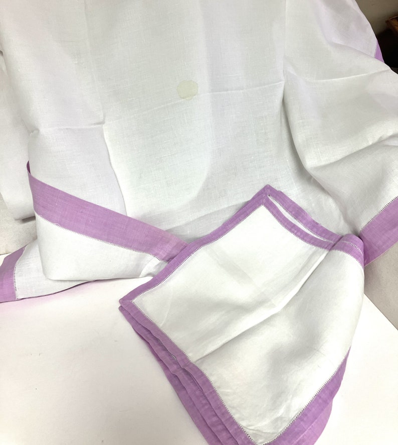 May include: White linen tablecloth with a lavender border and matching napkins. The tablecloth is folded, showing the lavender trim and a corner of a folded napkin. The fabric has a slightly textured appearance.