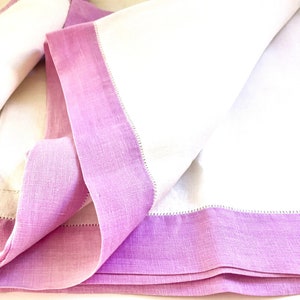 May include: White linen napkins with a lavender border. The napkins are folded, showcasing the contrast between the crisp white fabric and the soft, colored edge. The edges are finished with a delicate stitch.