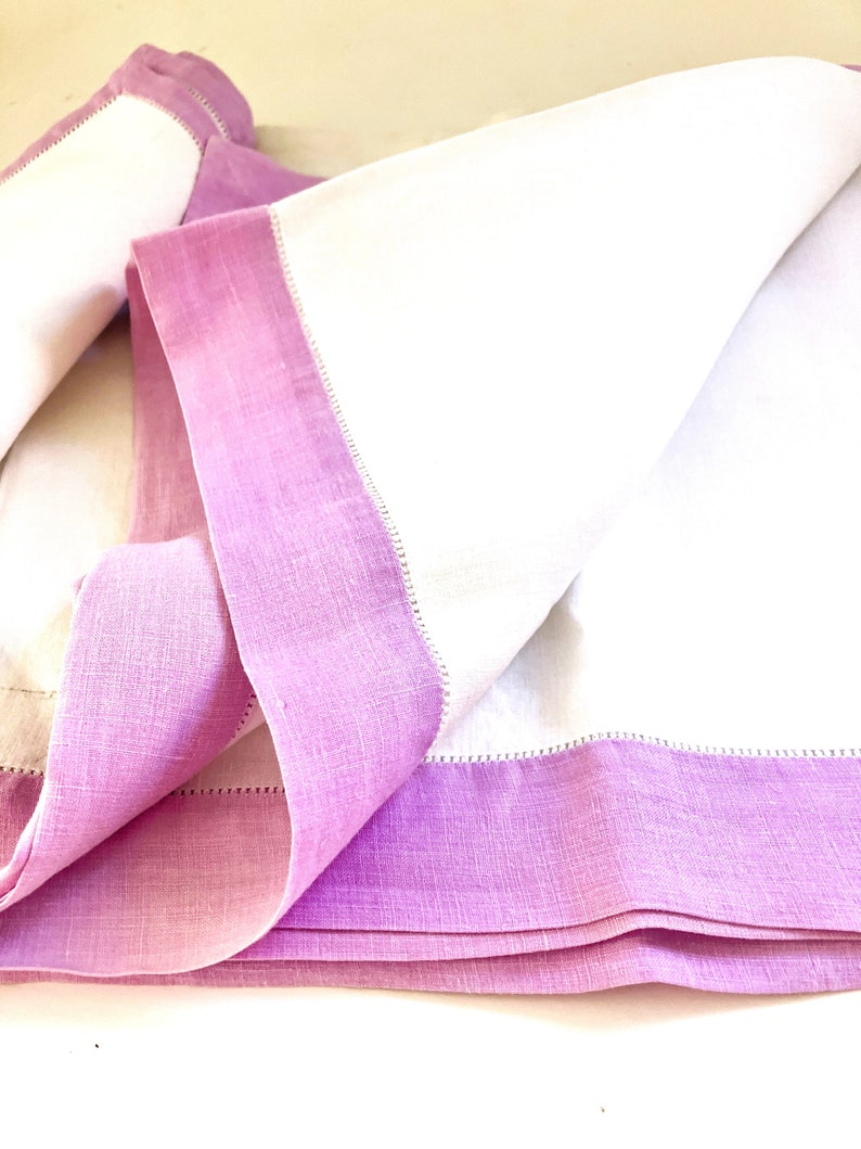 May include: White linen napkins with a lavender border. The napkins are folded, showcasing the contrast between the crisp white fabric and the soft, colored edge. The edges are finished with a delicate stitch.