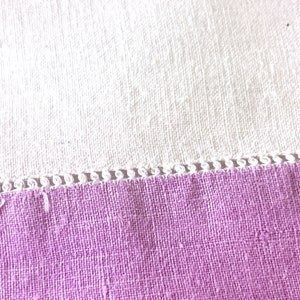 May include: Close-up of a lavender fabric with a white, textured fabric in the background. The lavender fabric has a decorative white trim along the edge. The fabrics appear to be linen or a similar woven material.