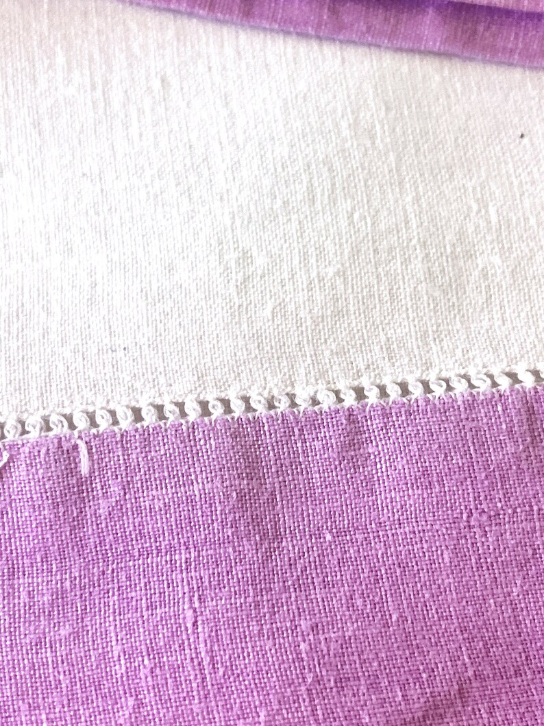 May include: Close-up of a lavender fabric with a white, textured fabric in the background. The lavender fabric has a decorative white trim along the edge. The fabrics appear to be linen or a similar woven material.