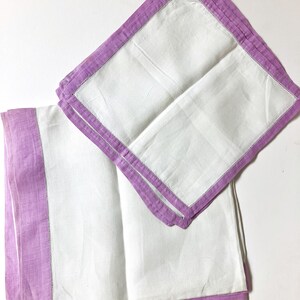 May include: Two white linen napkins with a lavender border and a decorative stitch. The napkins are folded and arranged on a white surface. The lavender border adds a pop of color to the otherwise simple design.