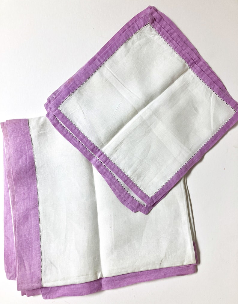 May include: Two white linen napkins with a lavender border and a decorative stitch. The napkins are folded and arranged on a white surface. The lavender border adds a pop of color to the otherwise simple design.