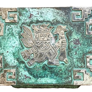 May include: Rectangular metal box with a silver-colored relief of a bird-like figure holding a staff. The box has a turquoise patina and silver-colored geometric border designs. The box has a hinged lid and is likely a decorative item.