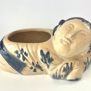 May include: A ceramic planter in the shape of a reclining figure. The planter is beige with dark blue accents and a serene facial expression. The open top is designed for planting small flowers or succulents.