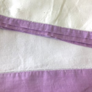 May include: Close-up of folded white fabric with a lavender border. The border is a solid color and is sewn with a decorative stitch. The fabric appears to be linen or a similar material.