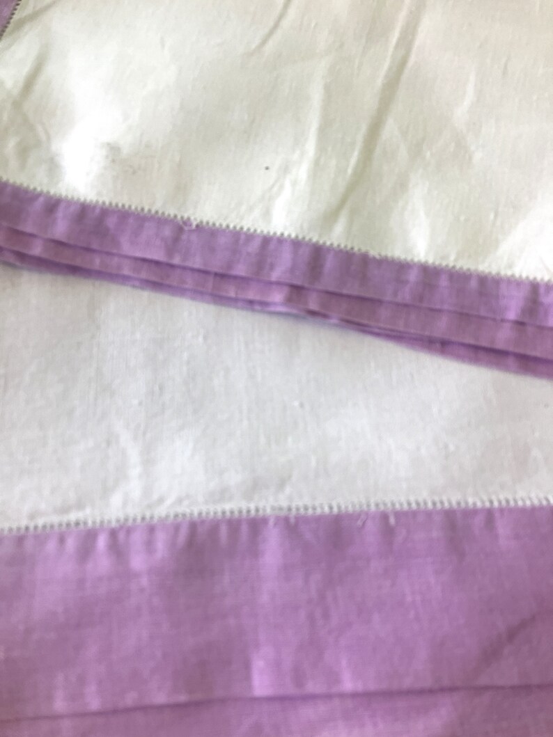 May include: Close-up of folded white fabric with a lavender border. The border is a solid color and is sewn with a decorative stitch. The fabric appears to be linen or a similar material.