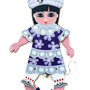 May include: A wooden doll with a painted design. The doll has a light blue hat with a floral design, black hair, and a purple dress with white floral accents. The doll's arms and legs are pink, and it has white pants with red and white accents. The doll's shoes are black with gray bows.