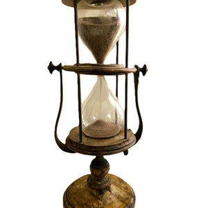 May include: A double-chambered hourglass featuring two clear glass bulbs connected by a central wooden frame. The structure includes a circular wooden base and top, supported by dark metal rods and curved side handles containing fine sand.