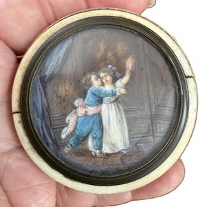 May include: A small, round antique miniature painting featuring two children in period clothing. The boy wears blue, the girl a white dress. The painting is set in a metal frame with an ivory-colored border. The children are embracing, with one holding a fan.