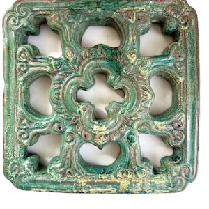 May include: Square, ornate architectural element with a distressed, aged appearance. The piece features a repeating openwork design in shades of green, red, and gold. The decorative piece has a weathered, vintage aesthetic.