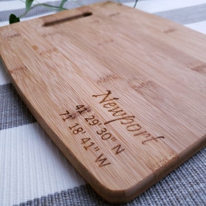 May include: A rectangular bamboo chopping board with a handle cutout. The board is engraved with the word "Newport" and coordinates 41° 29' 30" N, 71° 18' 41" W. The board has a natural wood grain.