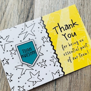 May include: A teal blue enamel pin with a white border and the text "TEAM AWESOME" in black. The pin is on a white card with a black and white star pattern. The card also has a yellow card attached to it with the text "Thank You for being an essential part of our team!" in black.