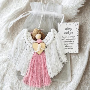 May include: A handcrafted macrame angel with white wings, a pink dress, and a wooden heart. The angel has a small tag that reads "Always with you". The angel is packaged in a sheer white bag.