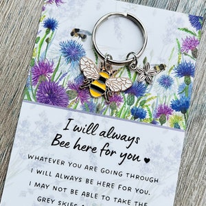 May include: A yellow and black bee charm attached to a silver keyring with a small white flower charm. The charm is on a card with the text "I will always Bee here for you" and a floral background.