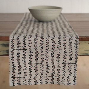 May include: A beige table runner with a repeating pattern of black vines, red berries, and small black birds. A light gray bowl sits on the runner. The runner is made of a natural-looking fabric.