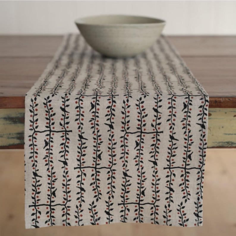 May include: A beige table runner with a repeating pattern of black vines, red berries, and small black birds. A light gray bowl sits on the runner. The runner is made of a natural-looking fabric.