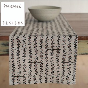 May include: A beige table runner with a repeating pattern of black birds and red berries on vines. The text "memi DESIGNS" is in a white box in the upper left corner. A light-colored bowl sits on the table runner.
